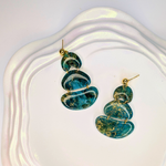 Handcrafted teal ocean-inspired earrings with gold flecks displayed on a white ceramic plate, featuring an organic oval shape and glossy resin finish